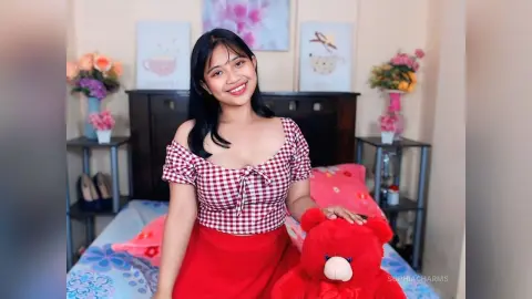 Smiling young woman in a red checkered top and red skirt sitting on a bed with a red teddy bear.