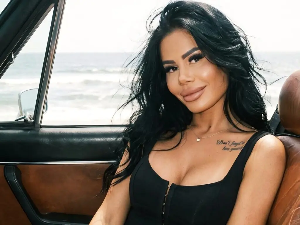 Smiling woman in a black top sitting in a vintage car by the ocean on a sunny day.