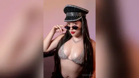 Young woman in a sparkly silver bikini and black captain's hat wearing sunglasses in a soft pink studio setting.