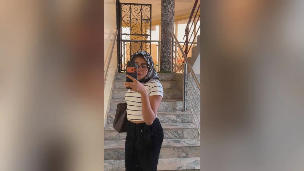AlbertNina — AlbertNina takes a mirror selfie on a staircase wearing a striped crop top, black pants, and a patterned headscarf.