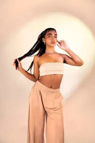 Veronica_Hottiee poses against a bright light, wearing a tan tube top and matching wide-leg trousers while holding her long black hair.