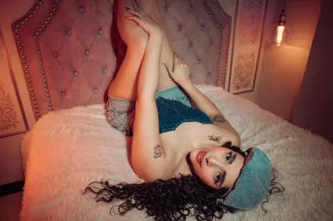 Young woman in denim shorts and top relaxing on a white faux fur bed in a warm, moody bedroom setting.