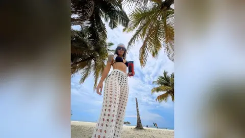 AlesiaNoir — Stylish woman in white crochet wide-leg pants and black bikini top posing under palm trees on a sunny beach.