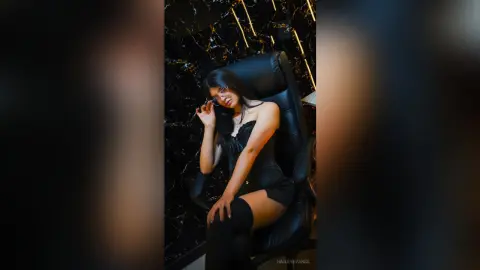 Elegant woman in a black cocktail dress and glasses sitting in a luxury dark interior setting.