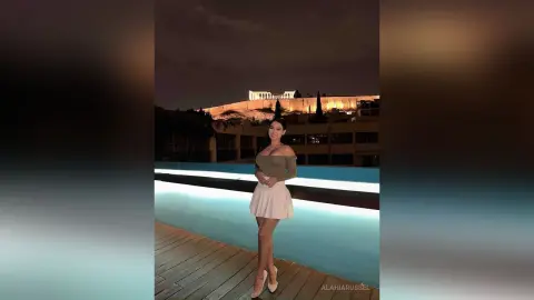 Smiling woman in a green top and white skirt standing by a pool with the illuminated Acropolis at night.