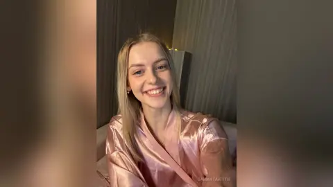 Smiling blonde woman in a pink satin pajama robe relaxing in a cozy, dimly lit bedroom.