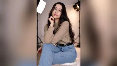 Smiling young woman in a beige sweater and blue jeans sitting in a professional photo studio.