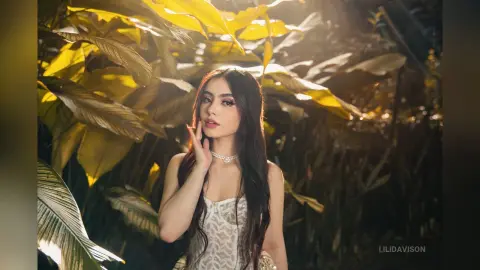 Young woman in a white lace top posing amidst lush tropical leaves in warm, golden sunlight.