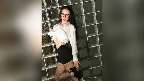 Stylish woman in white button-down shirt and black skirt posing confidently against a glass block wall.