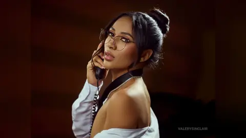 ValerySinclair wearing glasses and a white shirt, posing with a telephone against a dark background.