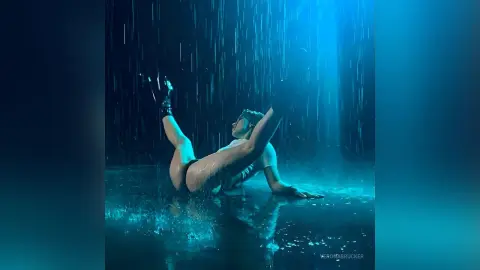 VeronaBrucker poses naked in the rain, arching her back while lying on a wet surface under blue light.