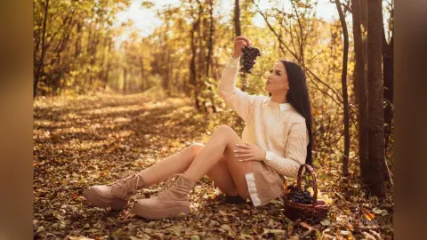 Woman in cream sweater sitting in autumn forest eating grapes from a basket amidst fallen leaves.