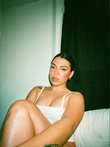 Young woman in a white tank top relaxing on a bed in a dimly lit room with a moody aesthetic.