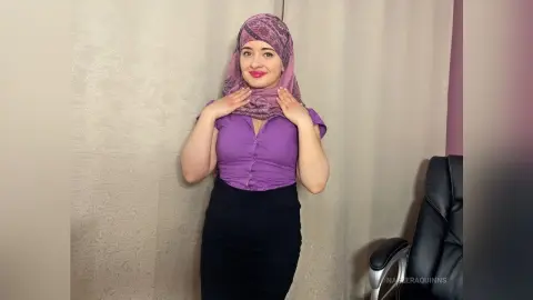 Smiling woman in a purple blouse and pink hijab standing against a neutral textured wall.