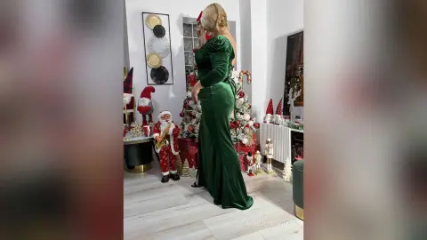 Elegant woman in a long green velvet gown standing in a festive room decorated with Christmas decor and Santa figures.