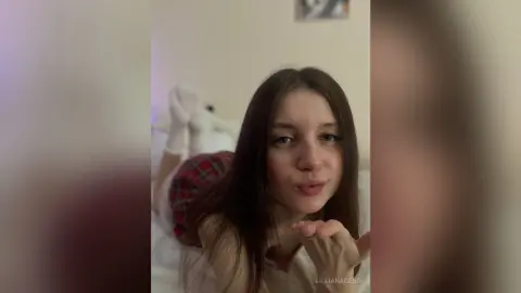 Young woman in red plaid shorts and white socks blowing a kiss in a cozy bedroom setting.