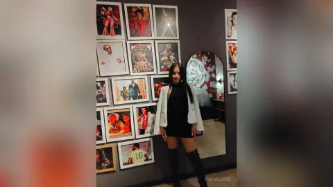 EvieMillers stands in front of a wall of framed photos, wearing a black dress, a white jacket, and long black boots.