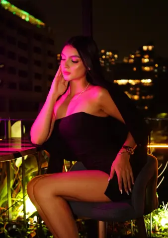 Elegant woman in a black strapless dress sitting on a rooftop terrace at night with city lights background.