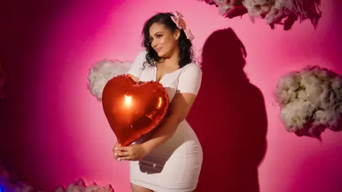 Woman in white dress holding a red heart balloon against a pink background for a romantic Valentine's Day mood.