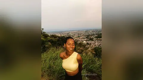 Smiling young woman in a yellow crop top enjoying a scenic view from a grassy hilltop at sunset.