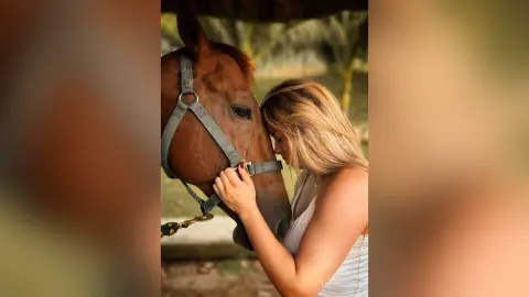 A blonde woman in a white tank top gently leaning her head against a brown horse in a peaceful outdoor setting.