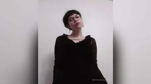 Young woman with short dark hair and glasses wearing a black blouse in a minimalist indoor setting.