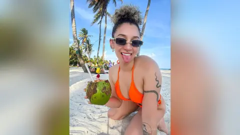 Smiling woman in orange bikini holding a coconut drink on a sunny tropical beach.