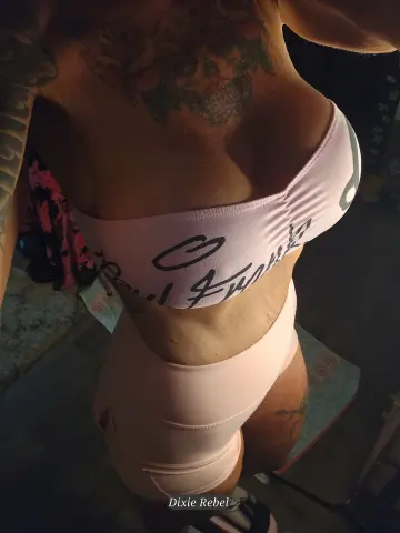 DixieRebelDixieDime — Tattooed woman in a pink sports bra and shorts posing in a dimly lit room.