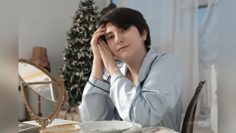 Young woman in light blue silk pajamas relaxing by a Christmas tree in a bright, cozy room.