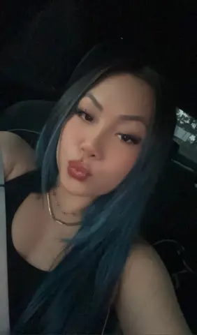 Young woman with long blue hair in a black tank top making a pouty face inside a car at night.