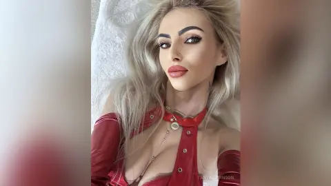 Blonde woman in a red satin outfit posing with glamorous makeup in a bright, soft-lit setting.