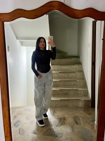 Young woman in a black long-sleeve top and grey trousers taking a mirror selfie in a modern home hallway.