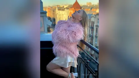 StellaFletcher poses on a balcony wearing a fluffy pink faux-fur top and a white satin skirt.