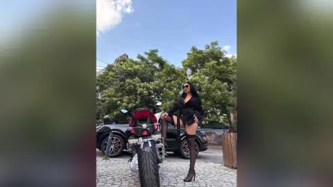 NicoleBella stands next to a black car and a motorcycle, wearing a black top, black shorts, and black thigh-high stockings with black heels.