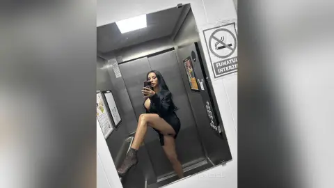 NicoleBella poses in a mirror selfie inside an elevator, wearing a black dress and black mesh stockings.