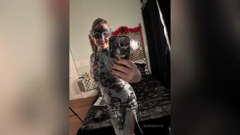 Smiling woman in a silver sequin cocktail dress and masquerade mask taking a mirror selfie in a bedroom.