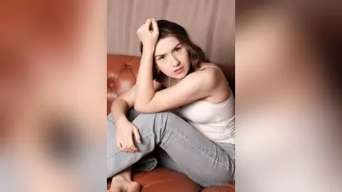 Young woman in a white tank top and light blue jeans sitting on a brown leather sofa with a thoughtful mood.