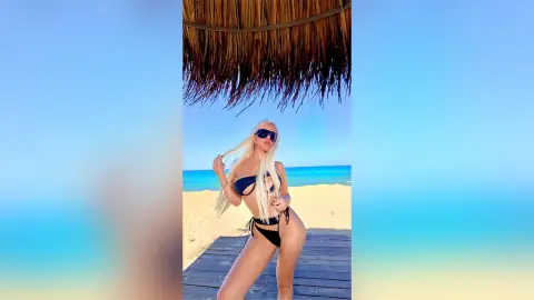 Blonde woman in a black bikini posing under a straw beach umbrella on a sunny tropical beach.
