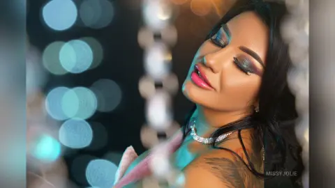 MissyJolie poses with eyes closed, wearing heavy eye makeup and a silver necklace against a soft, blurred background.