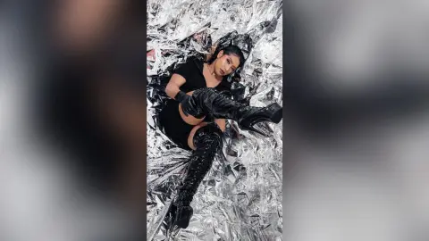 KimRoberth lies back on a shiny silver foil background wearing a black dress and thigh-high black boots.