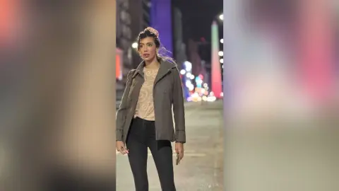 HaleyLyon stands outdoors at night wearing a grey jacket over a light tan top and black pants, with her hair pulled back and pink flowers in it.