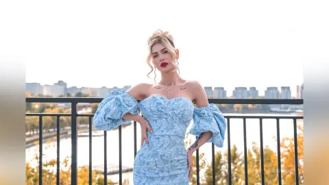 DaisyLin poses on a balcony wearing a light blue off-the-shoulder lace dress with blonde hair styled in a bun.