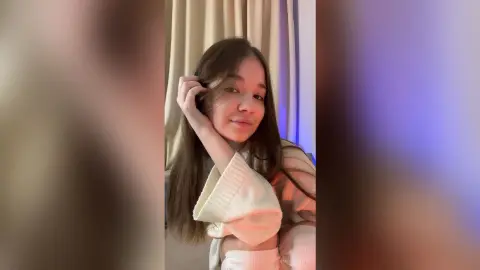 EmorySwift poses with her hand in her long brown hair, wearing a white long-sleeved sweater against a neutral curtain background.