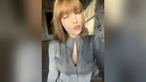 SharikaGlidden wearing a tight grey long-sleeve top with a deep zipper opening, showing her cleavage, with her light brown hair and bangs.