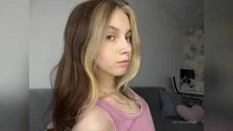 Young woman with long blonde hair wearing a pink ribbed tank top in a cozy indoor setting.