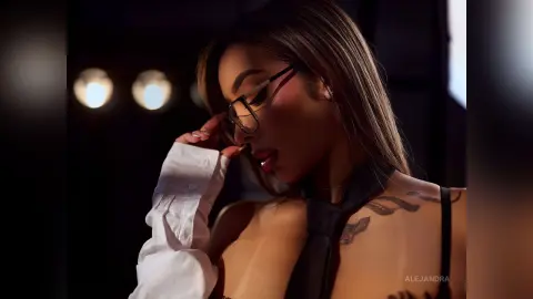 Stylish woman in a white shirt and black tie with tattoos posing in moody, dim studio lighting.