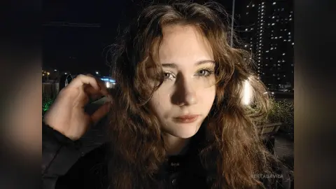 Young woman with wavy brown hair in a black jacket posing outdoors at night near city lights.
