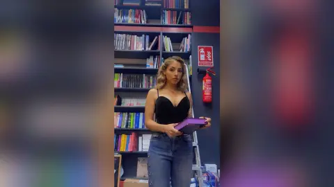 Young woman in a black bodysuit holding a purple book in a library setting.