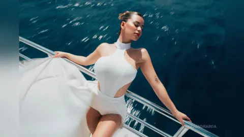 Elegant woman in a flowing white halter dress posing on a boat against deep blue ocean waves.