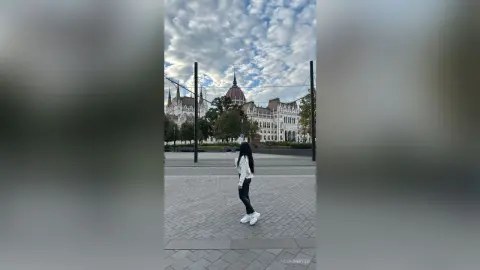 Young woman in white shirt and black pants standing in a city plaza near historic architecture under a cloudy sky.
